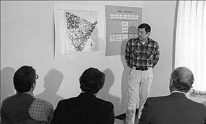 An Asian man stands beside a a map and chart in front of three seated men whose backs are to the camera. 